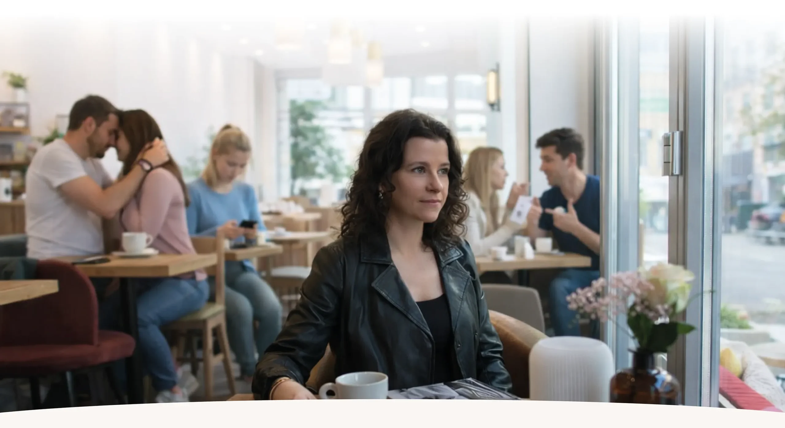 Nina Hömmen Single- und Beziehungscoaching Nina Hömmen sitzt nachdenklich in einem Café in Oldenburg. Im Hintergrund sind Paare zu sehen – einige liebevoll verbunden, andere im Streit – sowie eine Frau, die allein auf ihr Handy schaut. Die Szene symbolisiert Beziehungsmuster, Einsamkeit, Nähe und die unterschiedlichen Facetten von Liebe und Verbindung.